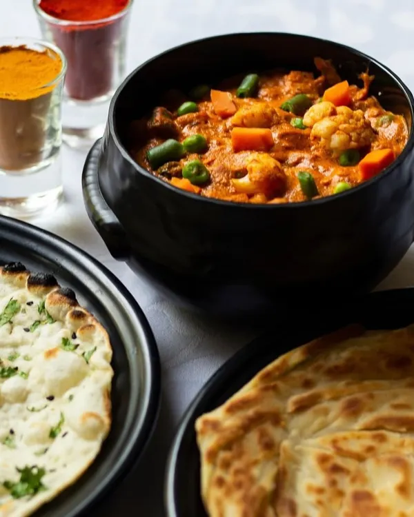 A wholesome North Indian meal featuring a rich vegetable curry, served with soft naan and flaky paratha. The small glasses contain the essential spices that give our curries their authentic flavor.