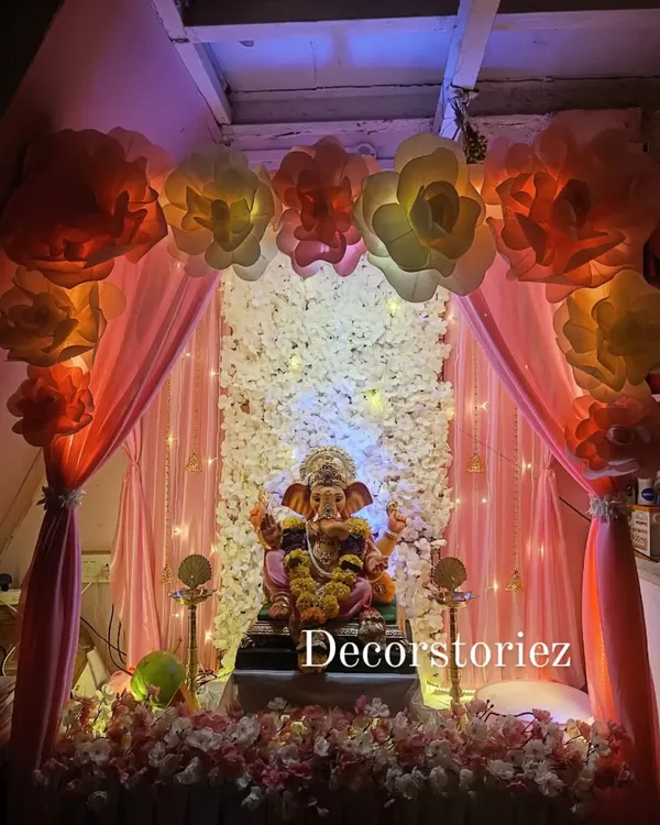We designed this grand and elegant home setup for the Ganpati festival. The backdrop features a wall of white artificial flowers, soft peach draping, and oversized paper flowers, all illuminated to create a divine atmosphere for Bappa.