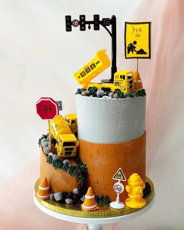 A two-tiered construction-themed cake with incredible detail. It features a winding road with toy trucks, traffic signs, and a "Dig In" sign, all on a cake designed to look like concrete and brick.