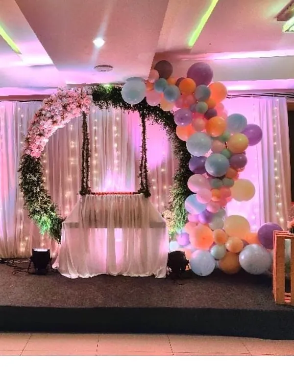 An ethereal cradle ceremony setup featuring a floral wreath and a cascade of pastel balloons. The soft lighting and elegant swing create a perfect, serene atmosphere for this special occasion.