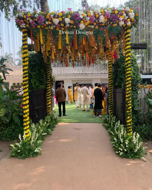 A festive entrance for a Mehndi ceremony. The arch is decorated with yellow and white flowers in a spiral pattern, with traditional marigold tassels hanging from the top to welcome guests.