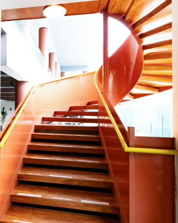 The striking spiral staircase at the Sona Vistaas Clubhouse, seen from a lower angle. Its continuous, flowing form in a bold terracotta red acts as a sculptural centerpiece, seamlessly blending movement, structure, and visual drama within the multi-level space.