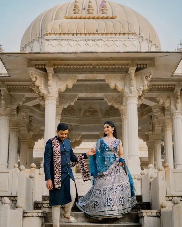 A classic shot of a couple descending the steps of a marble cenotaph at Gaitor Ki Chhatriyan. The groom gently leads the bride, showcasing the intricate details of their traditional blue outfits against the white marble.