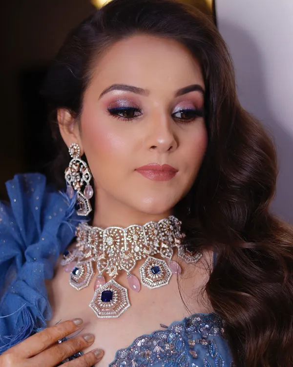 A close-up of a stunning cocktail look. The focus is on the shimmery blue and purple smokey eye, which beautifully complements the client's blue outfit and diamond jewelry.