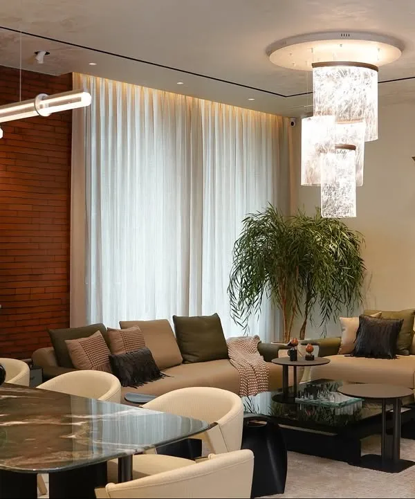 A complete view of the living and dining area at our Amaryllis project, showcasing the layered lighting from the sculptural chandelier and linear pendant. The space is unified by earthy tones and an exposed brick wall, creating a warm, cohesive environment.