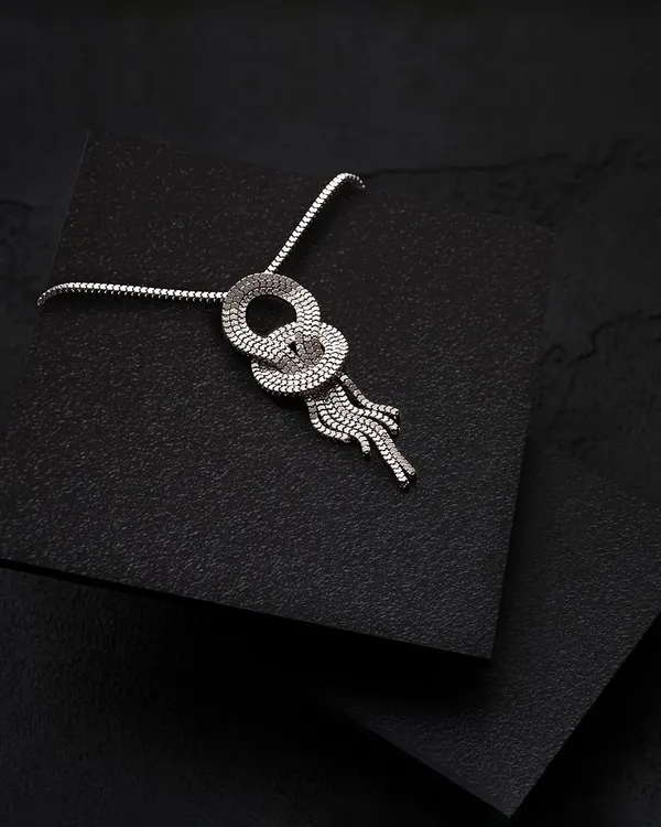 An elegant silver necklace with a knotted design, photographed on a textured black surface. The minimalist setup and focused lighting draw all attention to the product's unique form and craftsmanship.