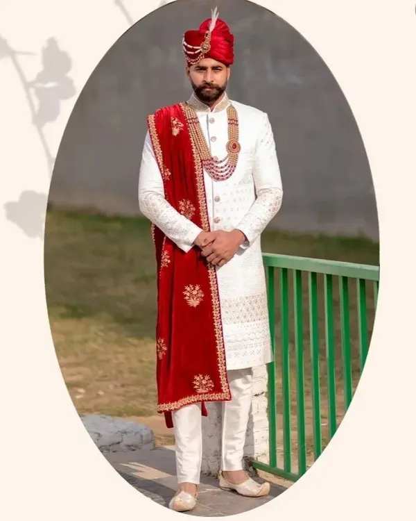 A groom looking sharp in a classic ivory sherwani paired with a rich red velvet stole. The intricate embroidery and matching red pagri create a regal and coordinated look.