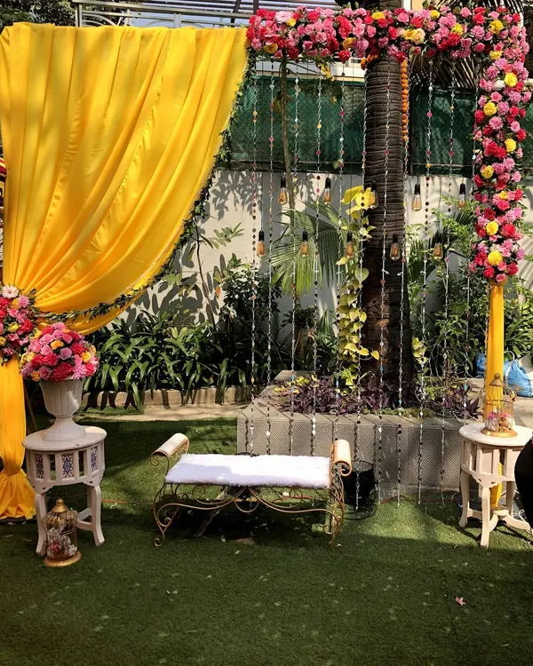 A bright and cheerful photo booth designed for a Haldi ceremony. The setup includes a comfortable bench set against a backdrop of vibrant yellow drapes, floral arrangements, and hanging lights.