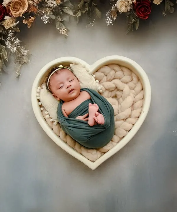 An overhead shot of a newborn nestled in a heart-shaped bowl, framed by an arch of beautiful flowers. This is a very popular and artistic newborn photography setup.