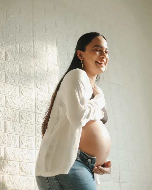 A joyful and candid lifestyle portrait. The mom-to-be is dressed in jeans and a simple white shirt, laughing naturally in the warm sunlight of our studio.