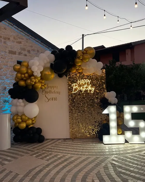 A sophisticated outdoor birthday setup featuring a modern white arch, a gold shimmer wall for photos, and a bold black, white, and gold organic balloon garland. The large, illuminated "15" marquee numbers and a custom neon "Happy Birthday" sign add a personalized and glamorous touch, perfect for milestone celebrations.