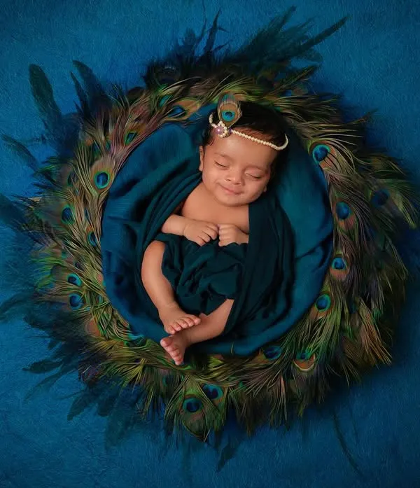 A peaceful portrait of a newborn styled as little Krishna, nestled in a bed of peacock feathers. This top-down shot highlights the full, artistic setup we create in our studio for themed newborn sessions.