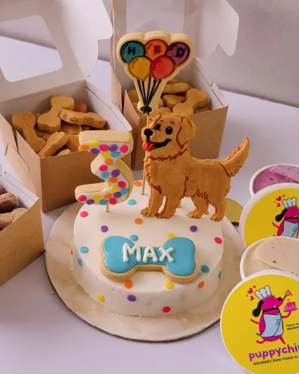 This is the ultimate birthday celebration package for a special doggo. It features a custom-decorated peanut butter and chicken cake with a golden retriever topper, personalised with the name 'Max', along with boxes of bone-shaped biscuits and individual pup-ice cream cups.