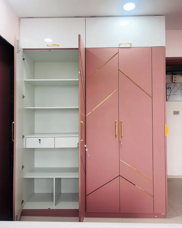 This pink and white hinged wardrobe showcases a clean, modern aesthetic with gold-accented geometric lines. The open door reveals the highly functional interior, complete with adjustable shelves, drawers, and dedicated compartments for organized storage.