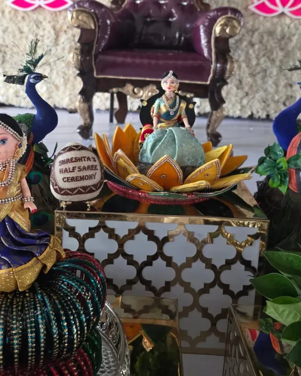 A close-up of the decorative elements for a half-saree ceremony, including traditionally dressed dolls, peacock figures, and a personalized coconut.