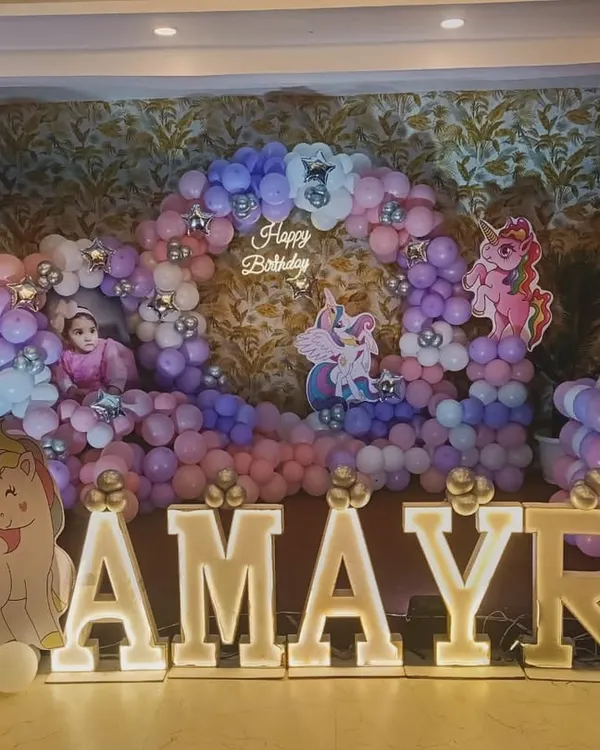 A magical unicorn-themed first birthday for Amayra. The decor features a large balloon ring in pastel pink and purple, unicorn cutouts, and big, bright marquee letters for the name.