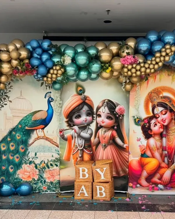 This vibrant baby shower backdrop features three distinct panels with illustrations of a peacock, baby Radha-Krishna, and Yashoda with Krishna. The scene is framed by a large balloon arch in metallic blue and gold, creating a grand and colorful setting.