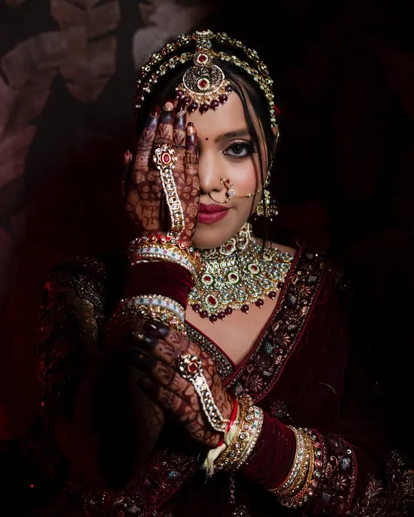A striking portrait of a North Indian bride, using her henna-covered hand to create a sense of mystery and draw attention to her eyes. The dramatic lighting highlights her beautiful jewelry.
