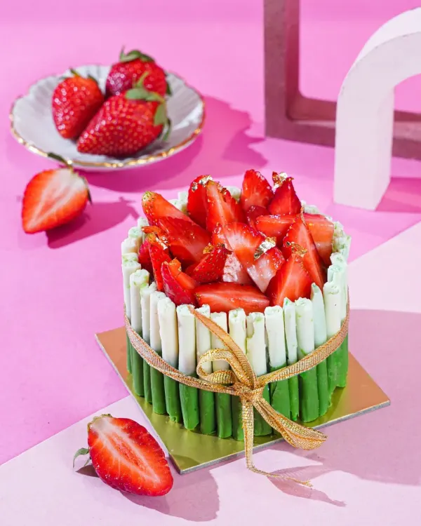 A beautiful strawberry cake with a green and white chocolate fence, tied with a golden ribbon. The bright pink background and modern props create a chic and celebratory feel.