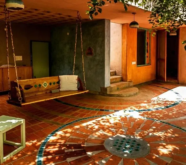 This sacred morning space in an artist's home is nestled amidst Honge trees. The design uses earthy lime plaster, terracotta floor tiles with a custom inlay, and a traditional jhula to create a shaded outdoor living area that feels both grounded and poetic.