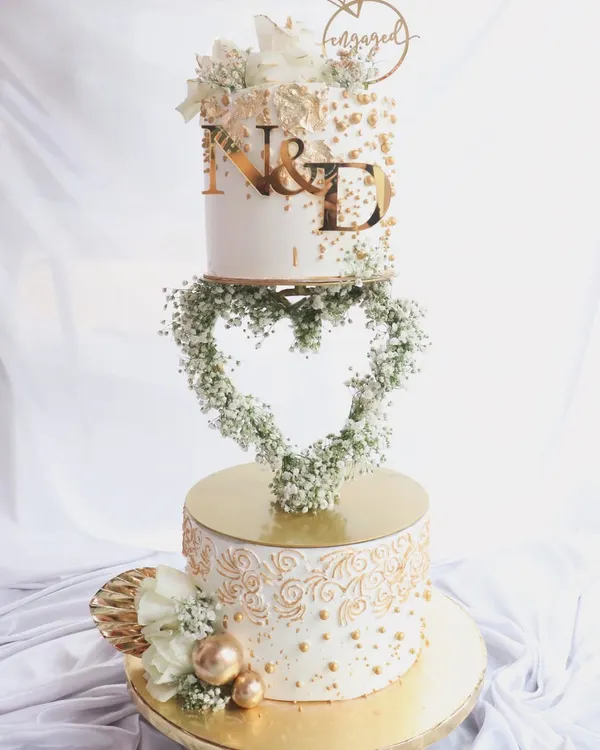This is one of my favorite engagement cakes, featuring a floating heart-shaped floral wreath. The two tiers are decorated with intricate gold piping and custom initials, making it a stunning centerpiece for a 'Baat Pakki' or engagement party.