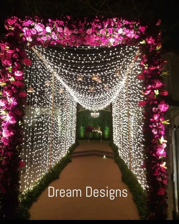 A gorgeous entrance tunnel for a grand wedding. The pathway is framed by an arch of purple roses and walls of cascading fairy lights, leading guests through a magical passage of light and flowers.