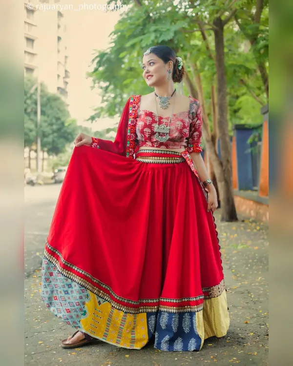 A full-length shot of a model in a vibrant red ghagra choli, taken outdoors on a city street. The image captures the flow of the skirt and the beautiful, multi-colored border.