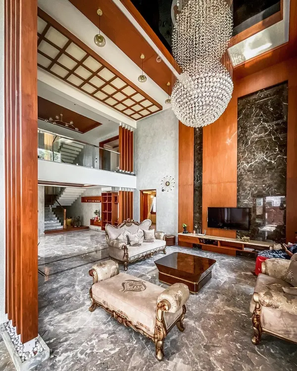 The grand living room of a private villa in Hennur, designed to impress with its double-height volume. A massive crystal chandelier, ornate furniture, and polished marble floors define this opulent space.