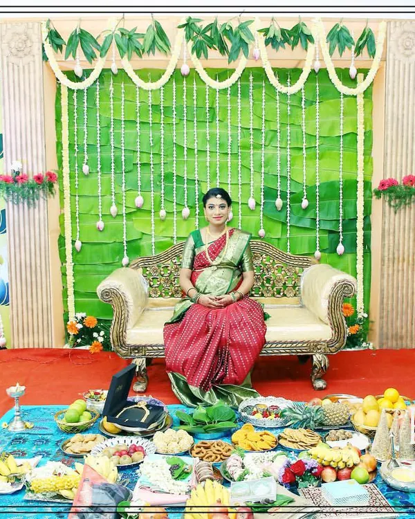 A beautiful stage setup for a Seemantham (baby shower) ceremony. The design features a backdrop of banana leaves and side panels with traditional lotus block prints, creating a natural and auspicious setting for the mother-to-be.