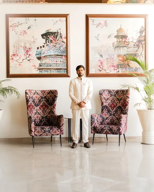 A full-length portrait of the groom, Piyush, in his elegant white engagement sherwani. The symmetrical composition with the large art pieces creates a formal and sophisticated look.