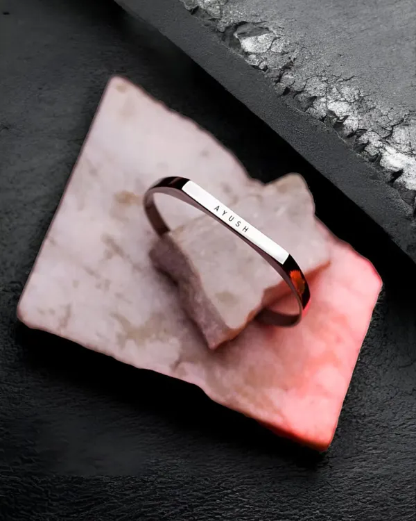 The black-plated cuff bracelet, engraved with 'AYUSH', resting on a piece of pink and white marble. This shot highlights the unique and modern appeal of the black finish.