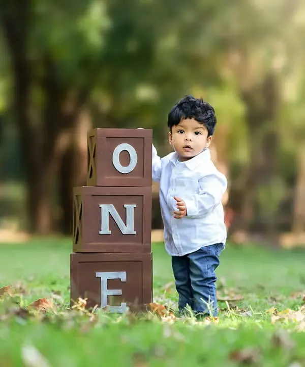 A classic outdoor portrait of a one-year-old boy standing proudly next to 'ONE' blocks in a lush green park setting.