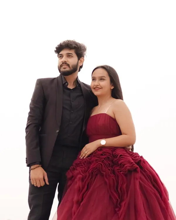 A classic and confident couple's portrait. The bride-to-be wears a stunning strapless maroon gown with intricate bodice detailing, perfectly complemented by her partner's sharp all-black suit.