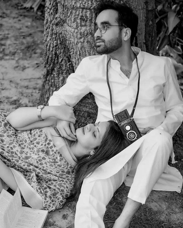 A beautiful black and white portrait capturing a quiet, romantic moment under a tree. The groom holds a vintage camera, and the couple's classic attire adds to the timeless, storybook feel of the image.
