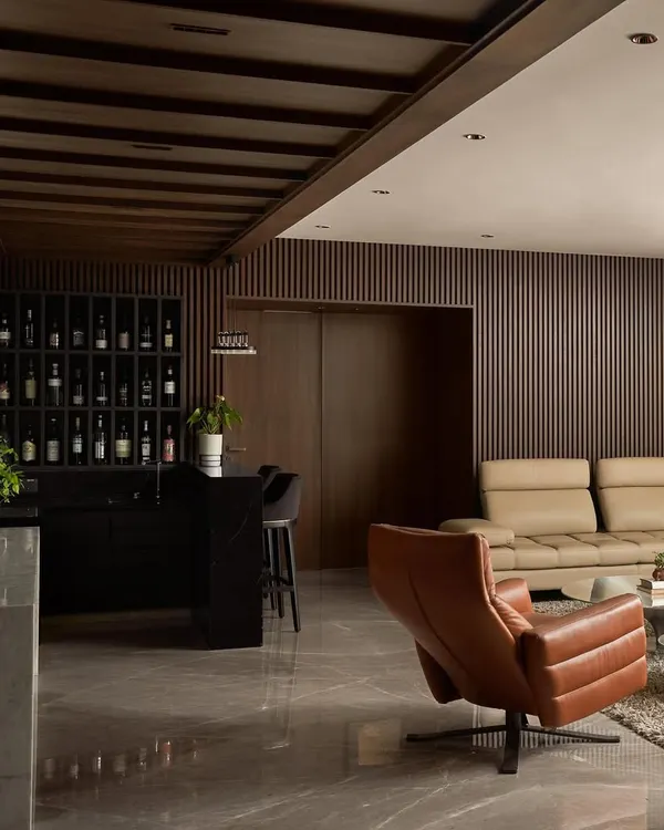 An overview of the Project Acropolis living room and bar area. The design uses a dark palette with fluted wood walls, leather furniture, and marble floors to create a sophisticated and cohesive environment.