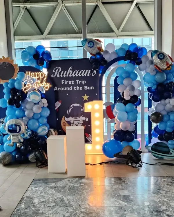 This outer space theme is one of my favorites for a first birthday party. The setup includes a full balloon arch in shades of blue, black, and silver, custom astronaut and rocket cutouts, and a large light-up number '1' to make the celebration for 'Ruhaan's First Trip Around the Sun' truly special.