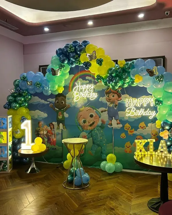 A large-scale Cocomelon birthday party setup. This decoration features a full backdrop, a vibrant balloon arch, multiple character cutouts, and two neon "Happy Birthday" signs.
