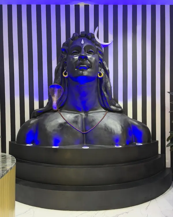 The final Adiyogi-inspired sculpture installed in the office lobby, illuminated with dramatic blue uplighting. The piece is set against a bold striped wall, creating a modern shrine that commands attention.
