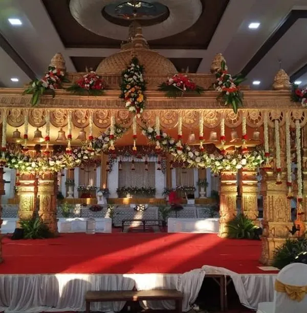 A beautiful and traditional marriage mantapa, featuring ornate golden pillars and a roof adorned with fresh flowers. The red carpet and stage create a classic and regal setting for the wedding rituals.