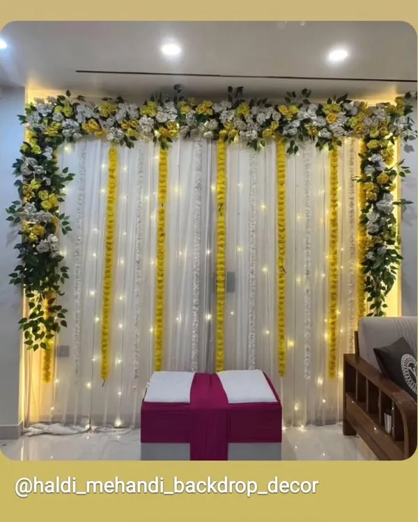 A simple and elegant indoor Haldi backdrop with white drapes, marigold strings, and fairy lights. The setup is completed with a floral border and seating, perfect for home ceremonies.