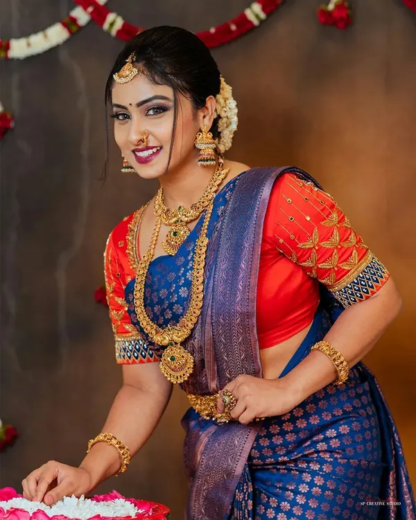 A classic studio portrait of a model in a royal blue silk saree with a contrasting embroidered red blouse. Her direct, smiling gaze and the clear visibility of the complete jewelry set make this a perfect image for a festive campaign.