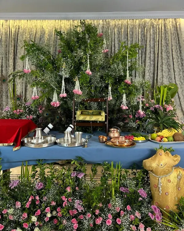 A beautiful Janmashtami altar ready for celebrations. The setup features a miniature swing for Lord Krishna, surrounded by lush greenery, pink flowers, and offerings of fruit, all set against an elegant curtained backdrop.