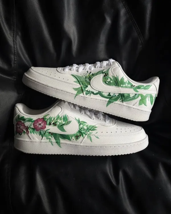 A pair of white Nike Court Vision sneakers adorned with a hand-painted design of fresh green leaves and pink flowers.