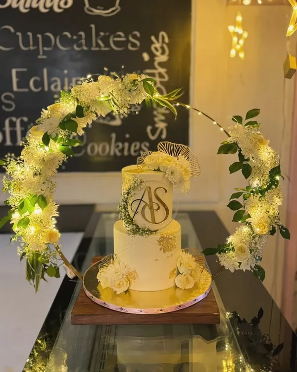 This two-tiered custom wedding cake features a stunning illuminated floral hoop, creating a breathtaking centerpiece for an evening celebration. The design incorporates a gold acrylic monogram, delicate white flowers, and soft lighting for a truly magical effect.