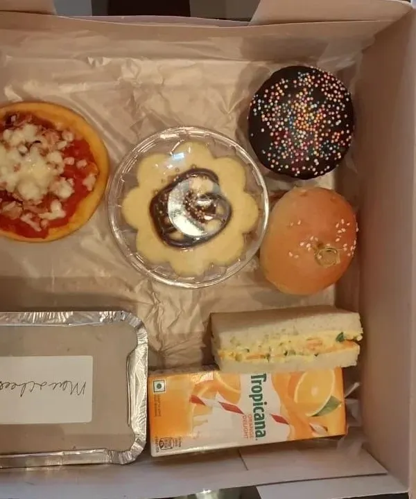 An overhead view of a packed kids' snack box. This assortment includes a mini cheese pizza, a chocolate cupcake with sprinkles, a sandwich, baked mac and cheese, and more to make any child's day special.