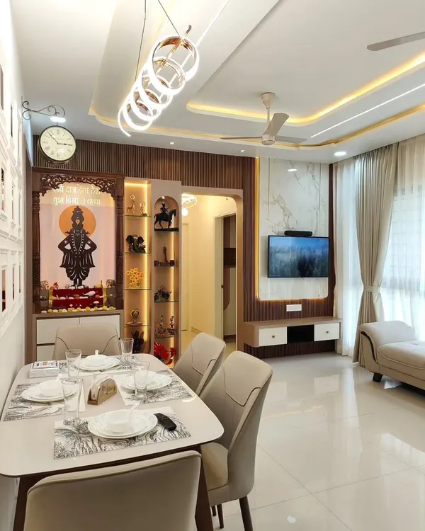 An open-plan living and dining area where the design flows seamlessly. The space includes a modern dining set under a statement chandelier, with the TV and pooja unit visible in the background, creating a unified look.