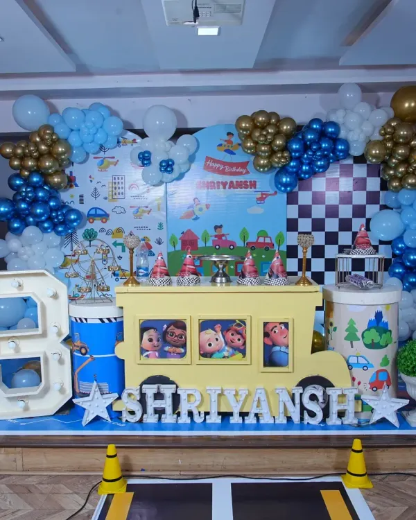This is a complete Cocomelon and transportation themed setup for a third birthday. It features a custom printed backdrop, a large balloon garland in blue, gold, and white, a light-up marquee number '3', and a school bus prop that doubles as a dessert table.