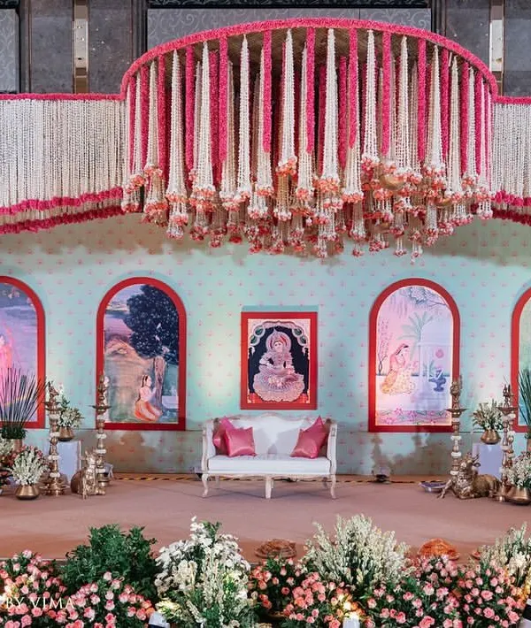 A grand and traditional stage for a South Indian half-saree function. The decor features massive floral chandeliers made of jasmine and pink flowers, with a custom backdrop depicting the stages of a woman's life.