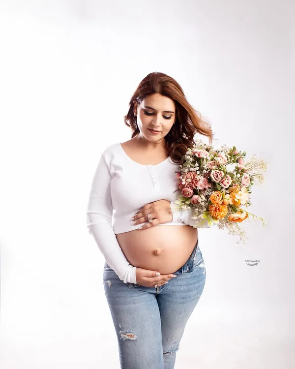 A beautiful and relaxed bare-bump portrait. The client wears a simple white top and ripped jeans, holding a bouquet for a fresh, natural, and modern maternity look.
