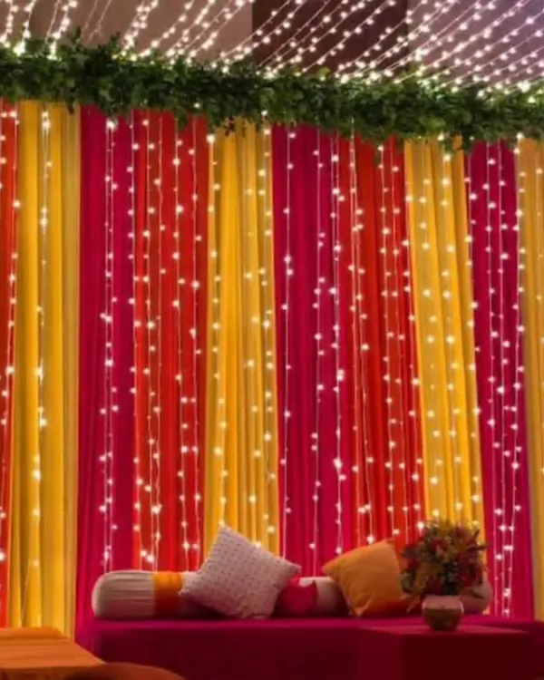 A vibrant backdrop for a Mehendi or Sangeet night, featuring colorful pink, yellow, and orange drapes. The entire setup is illuminated with strings of fairy lights for a magical effect.